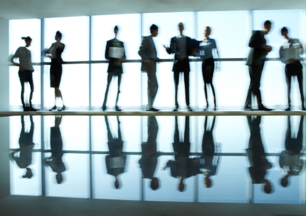 A group of business people standing in a hallway in front of a window. Their reflections are shown on the floor.